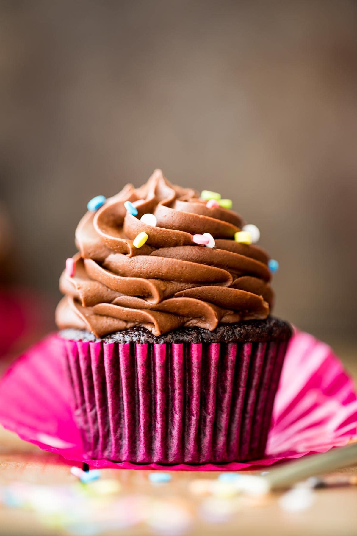 head-on view of a chocolate cupcake in a hot pink cupcake wrapper topped with piped chocolate frosting and rainbow sprinkles