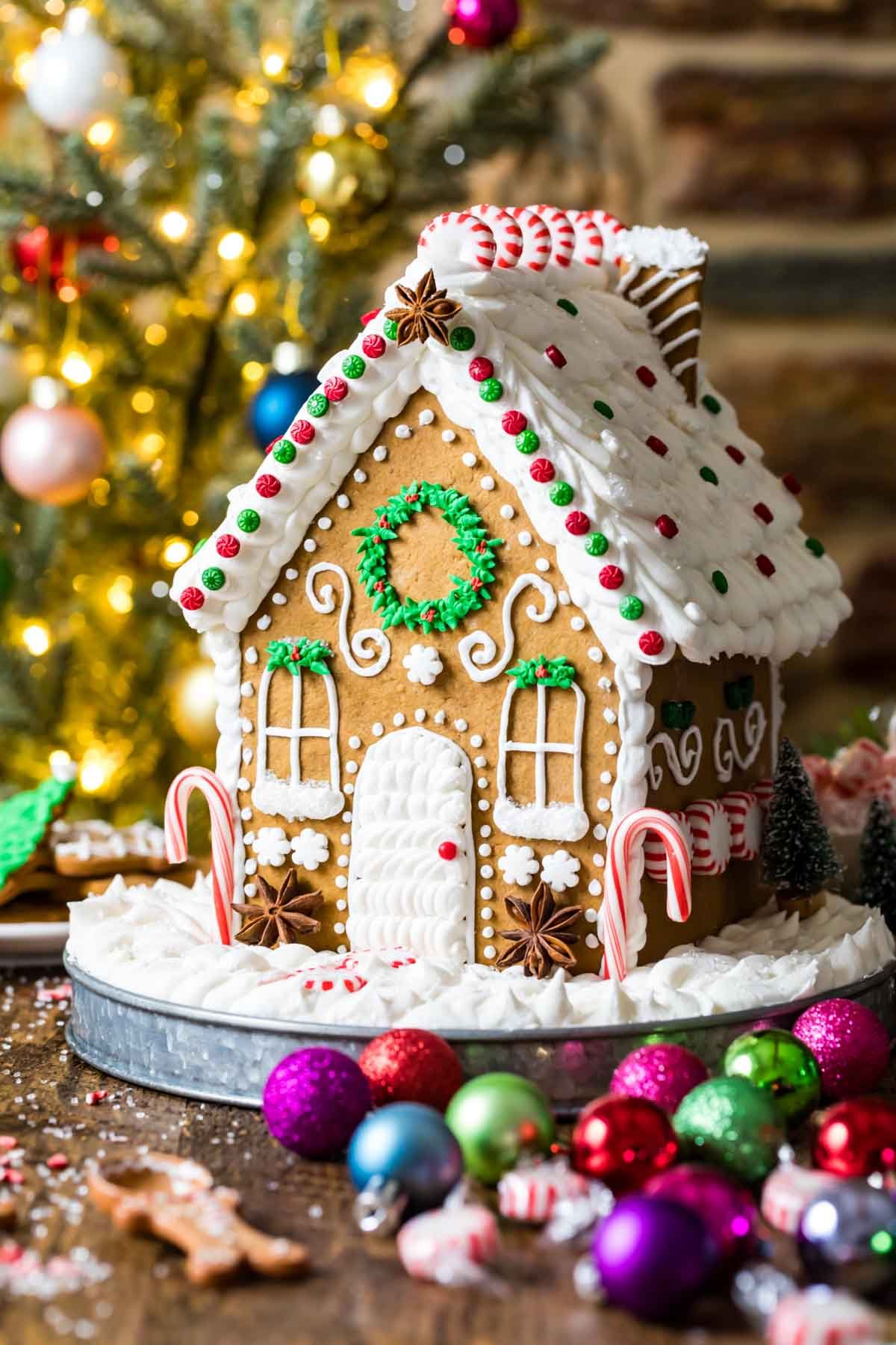 Intricately decorated gingerbread house on a metal plate with christmas ornaments in the foreground and a lit tree in the background.