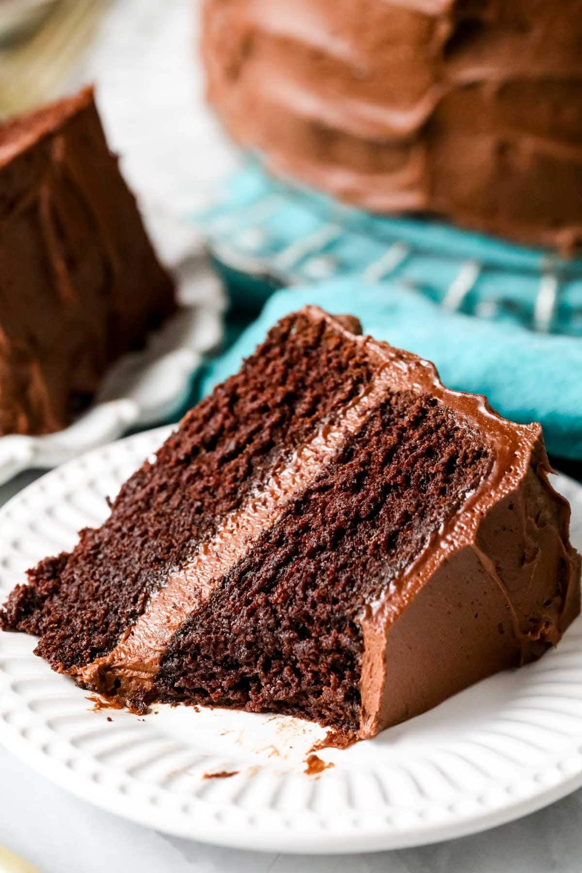 Slice of fudgy chocolate cake on a white plate with  cake on crystal platter over blue cloth in background.