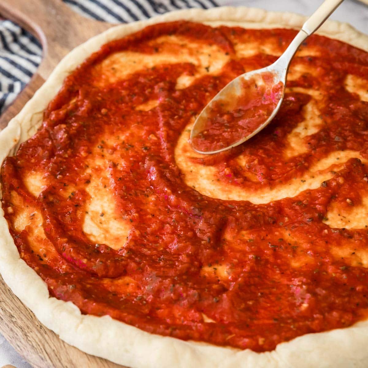 My rich red homemade pizza sauce recipe being spread onto an unbaked pizza crust.